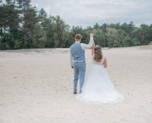 Yes! Getrouwd! Foto van een bruidspaar in Bergen op Zoom. Ze houden elkaars handen vast en steken één hand in de lucht. Trouwdag professioneel vastgelegd door trouwfotograaf Puur Geluk Fotografie.