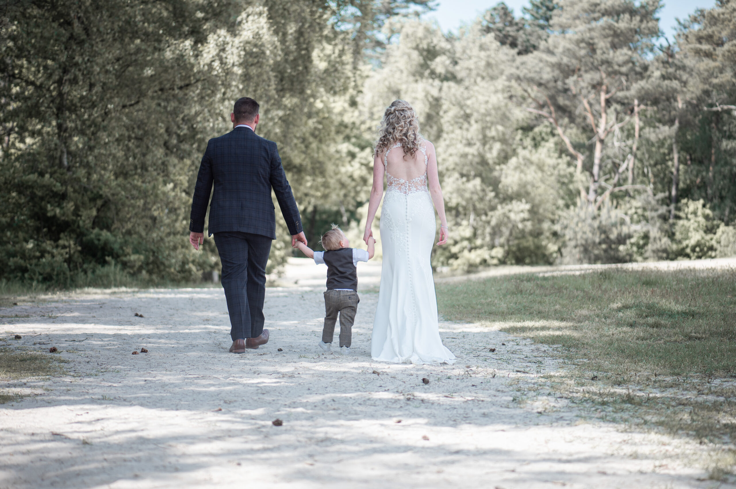 Bruidspaar met hun zoontje loopt hand in hand over een zandpad in het bos, lichte romantische fotografie vol verbondenheid. Foto door Puur Geluk Fotografie.
