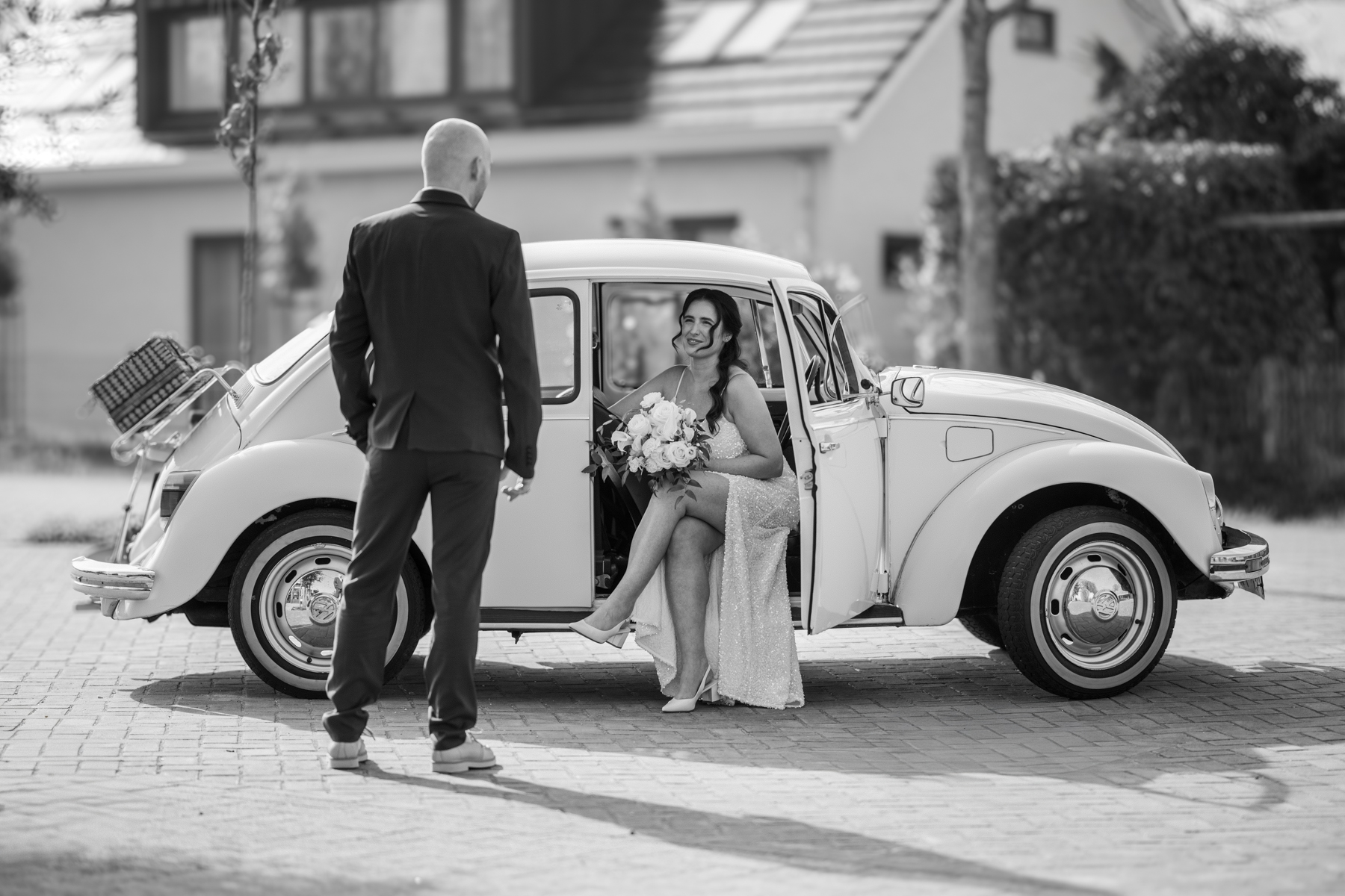 Puur Geluk Fotografie legt een bruid vast die glimlachend in de open deur van een klassieke Volkswagen Kever zit met een boeket in haar handen, terwijl de bruidegom haar tegemoet loopt in zacht, romantisch licht.